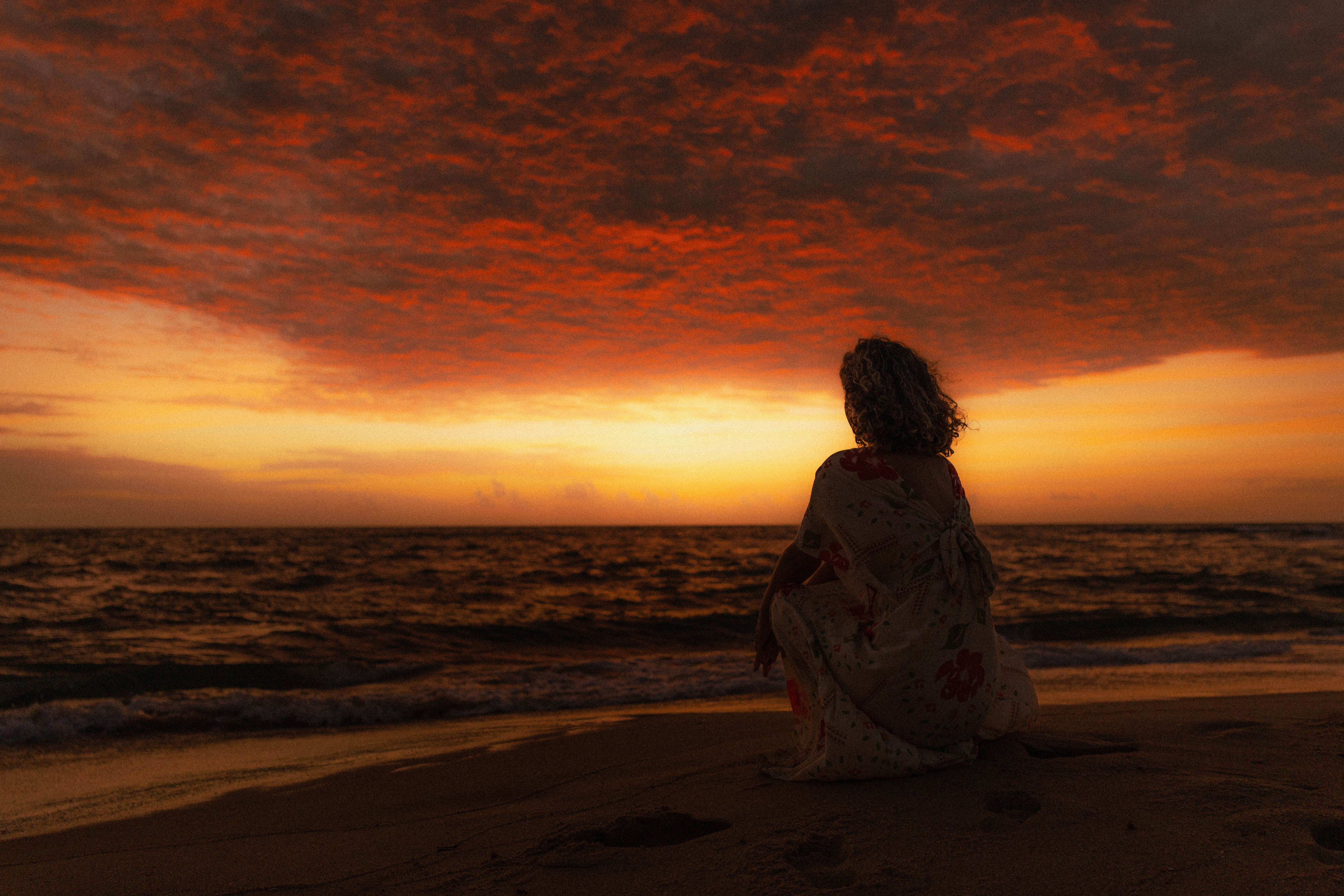 A woman sits quietly on a beach as the sky glows red and orange during sunset